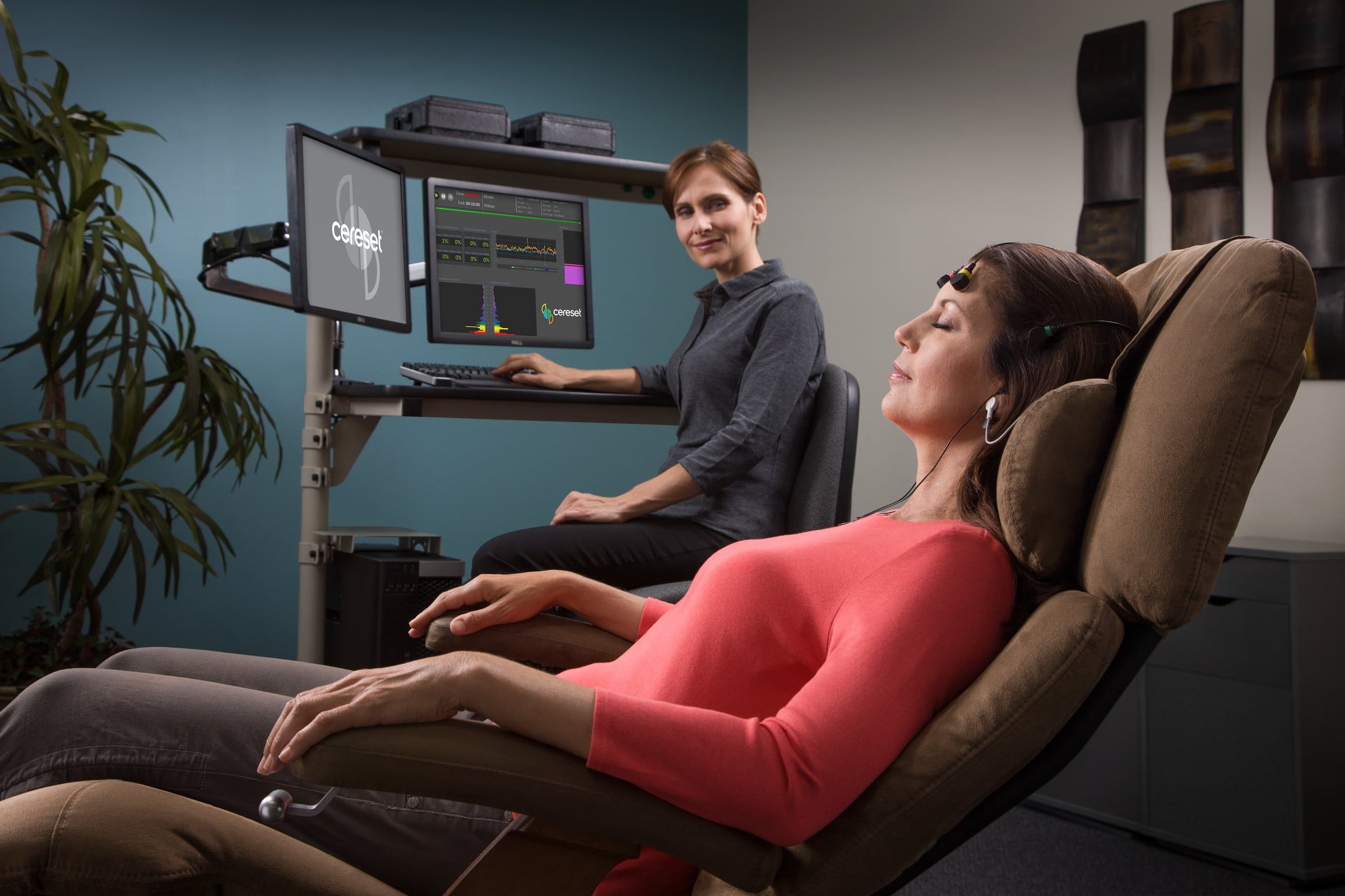 A client reclining with her eyes closed during a Cereset session, sensors and headphones in place, while a Cereset Tech Coach watches her brain activity on the monitors behind her